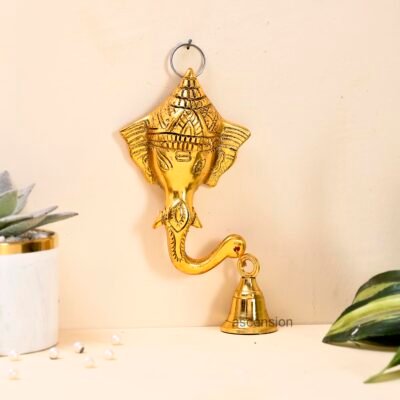 Golden brass Ganesha face wall hanging with a bell suspended from his trunk mounted on a beige wall