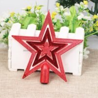 A red star Christmas tree topper with a hollow layered design resting against a white planter on a woven surface.
