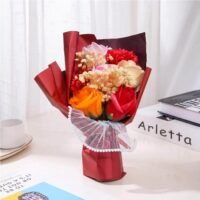 Ascension artificial rose flower bouquet in red wrapping next to a clear gift box printed with Best Wishes on a table.