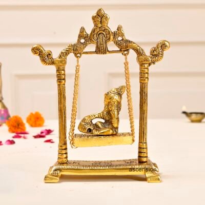 Golden brass Ganesha reclining on a swinging jhula suspended inside an arched metal frame on a white table