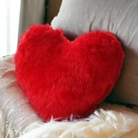 Fluffy red heart plush throw pillow leaning against a sofa cushion with a soft blanket.