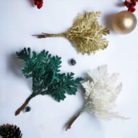 Three glitter artificial cedar sprigs in gold, green, and white resting on a white surface with a gold bauble and pinecone.