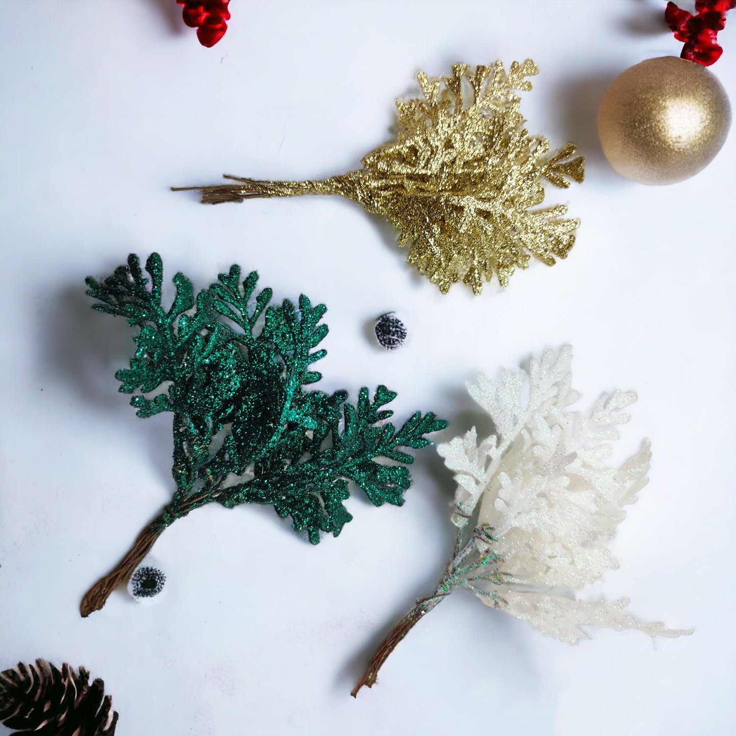 Three glitter artificial cedar sprigs in gold, green, and white resting on a white surface with a gold bauble and pinecone.
