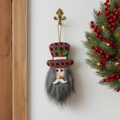 A plush Santa gnome ornament with a grey beard and patterned burgundy top hat hanging on a brass hook next to a Christmas wreath.