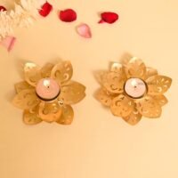 Two golden lotus tealight candle holders with burning tealights on a beige background with rose petals