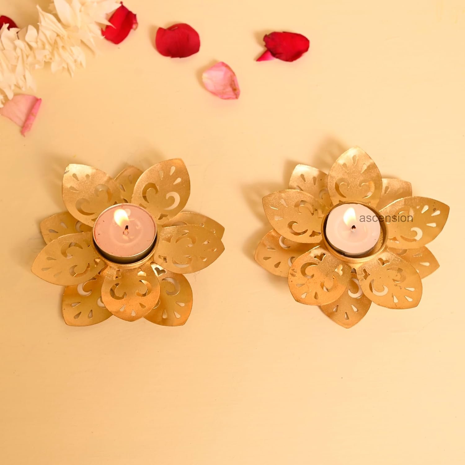 Two golden lotus tealight candle holders with burning tealights on a beige background with rose petals