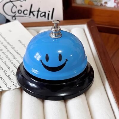 Bright blue metallic call bell with a black smiley face print and silver plunger on a black base.