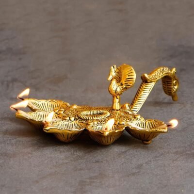 Golden brass peacock panch aarti diya with five lit wicks and a curved handle on a grey surface