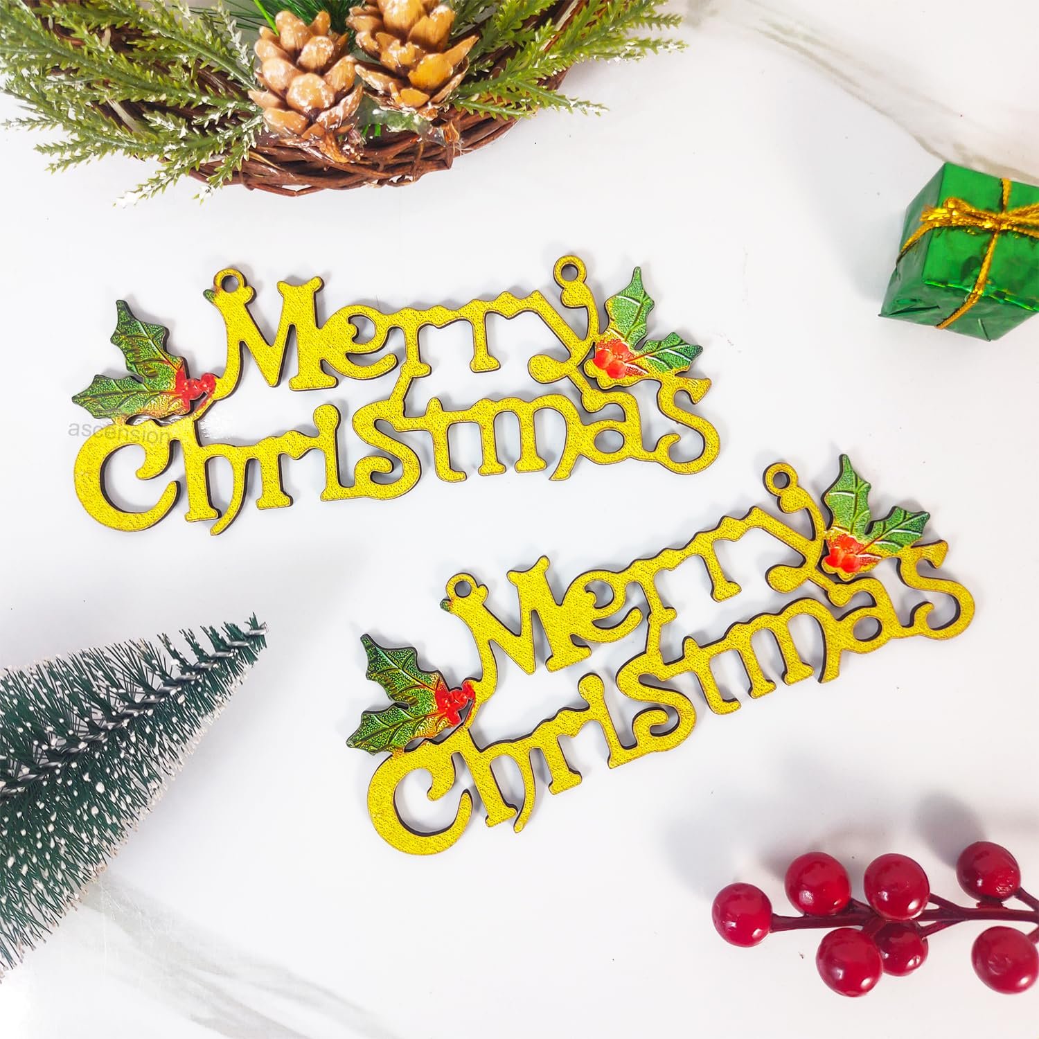 Two gold Merry Christmas signs decorated with green holly leaves and red berries resting on a white surface.