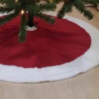 A red Christmas tree skirt with a white faux fur trim wrapped around the base of a lit pine tree on a wooden floor.