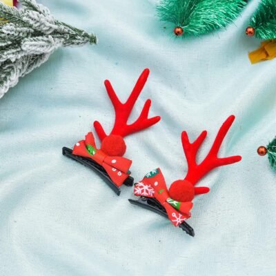A pair of red reindeer antler hair clips featuring festive bows and pom-poms resting on a light blue fabric.