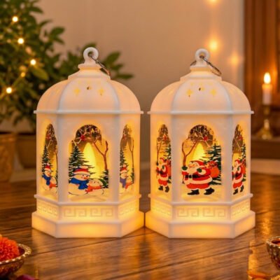 Two white decorative Christmas LED lanterns glowing on a wooden table, featuring colorful Santa Claus and snowman illustrations.