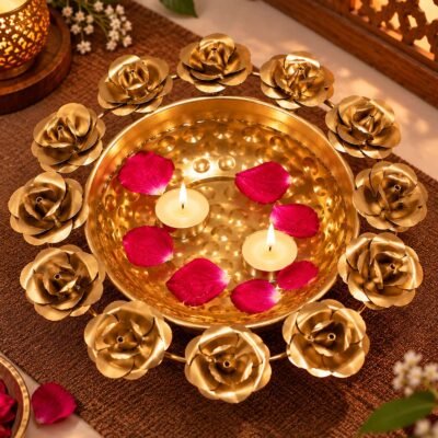 Overhead view of a golden brass urli bowl adorned with decorative metal roses, filled with water, red rose petals, and lit tealight candles.