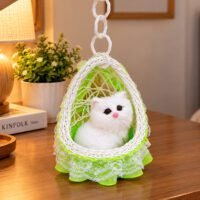 A realistic plush white cat figurine sits inside a hanging woven swing with green netting and lace trim on a wooden table.
