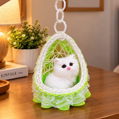 A realistic plush white cat figurine sits inside a hanging woven swing with green netting and lace trim on a wooden table.
