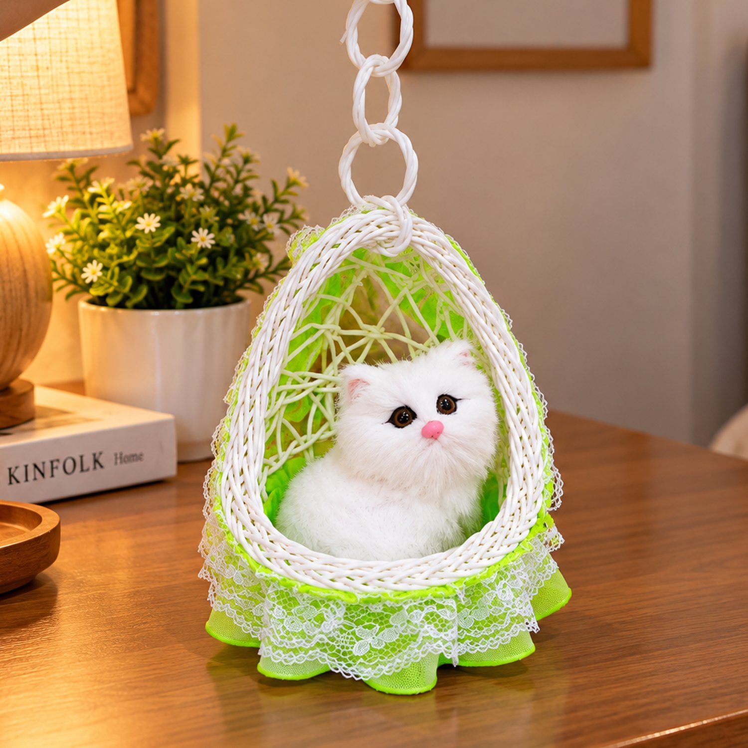 A realistic plush white cat figurine sits inside a hanging woven swing with green netting and lace trim on a wooden table.