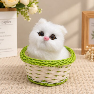 Realistic white plush kitten with brown eyes and a pink nose sitting in a green and beige woven basket.